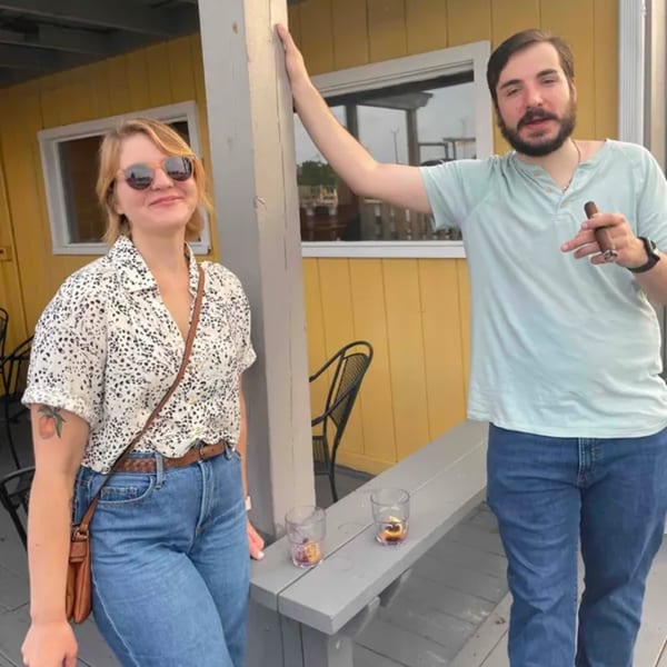 A man holding a cigar and a woman in sunglasses stand on a patio with drinks.