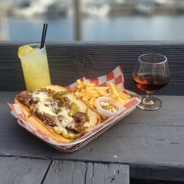Cheesesteak sandwich with fries, coleslaw, lemonade, and a glass of amber liquid on a wooden table.