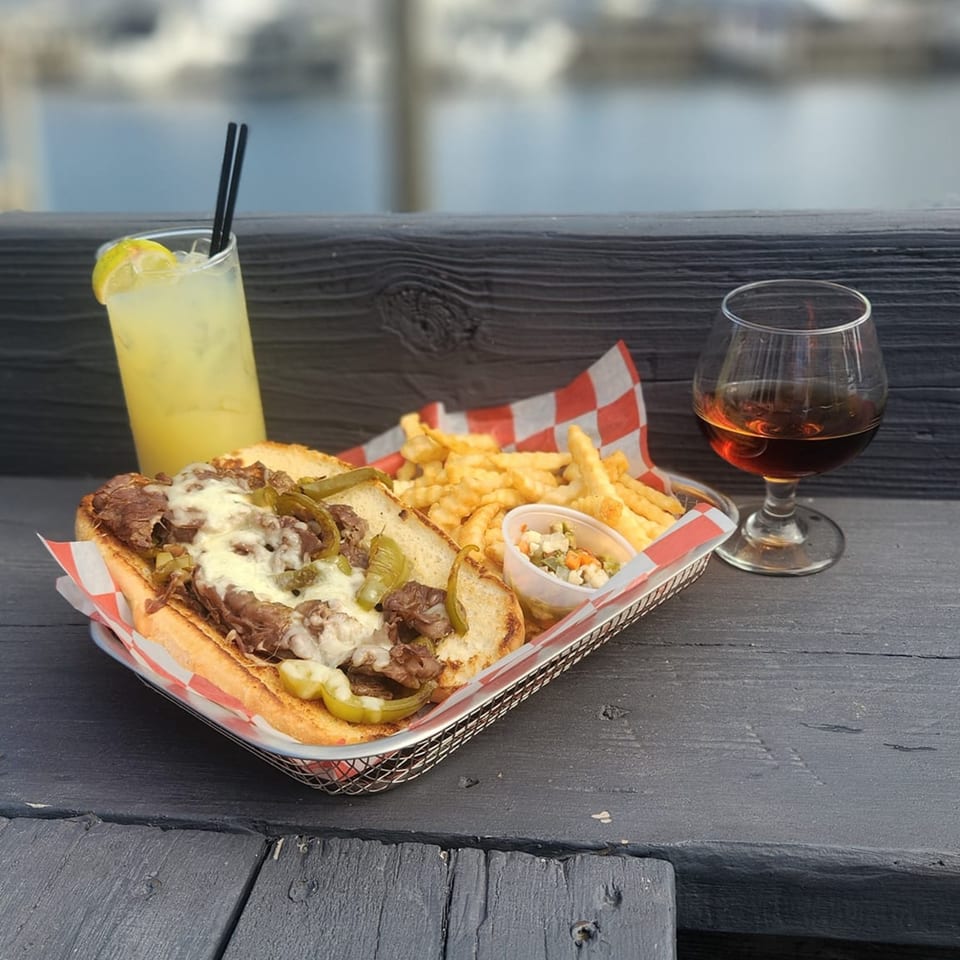 Cheesesteak sandwich with fries, coleslaw, lemonade, and a glass of amber liquid on a wooden table.