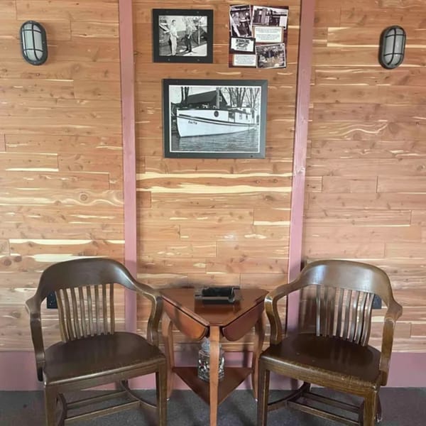 Two wooden chairs flank a small table against a wood-paneled wall with framed nautical photos.