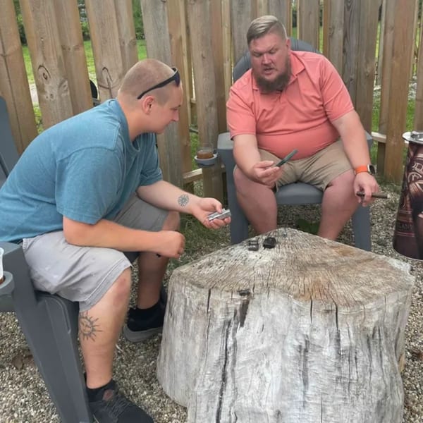 Two men sitting outdoors, one holding a cigar cutter, the other a cigar, with a tree stump table.