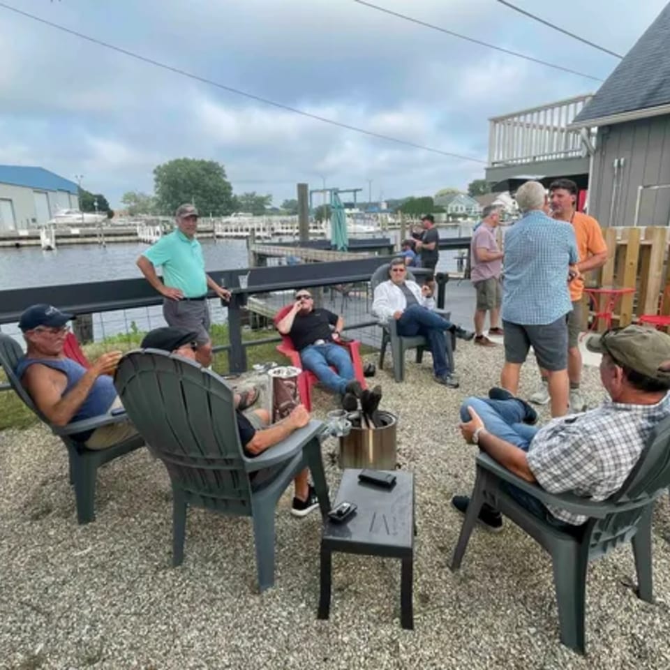 Men relaxing in chairs on a gravel patio overlooking a marina at On the River Tavern.