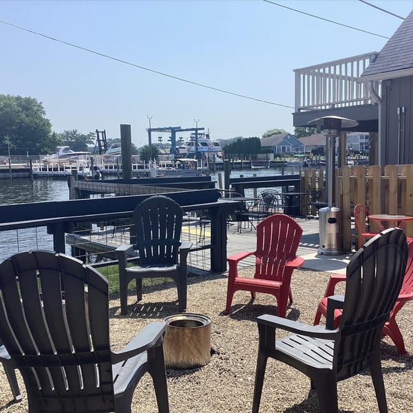 Outdoor seating area with red and black Adirondack chairs overlooking a river with boats.