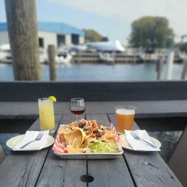 Nachos, drinks, and place settings on a wooden table overlooking a marina at On the River Tavern.