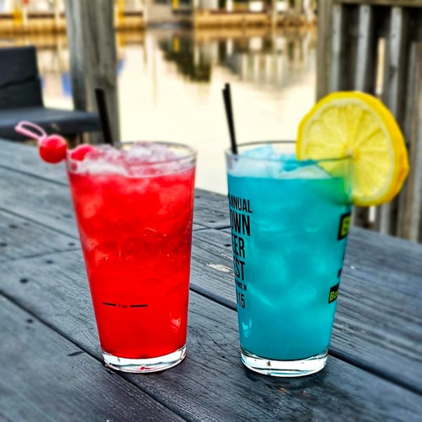 Two colorful cocktails, one red with cherries and one blue with a lemon, on a wooden table by the water.