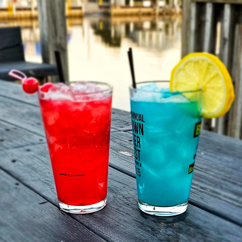 Two colorful cocktails, one red with cherries and one blue with a lemon, on a wooden table by the water.