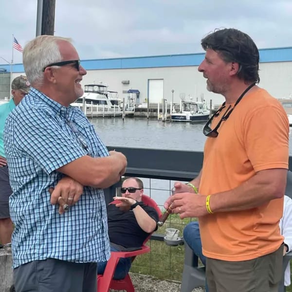 Two men talking and smiling outdoors by a marina, one holding a cigar.