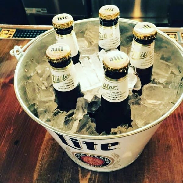 Five Miller Lite beer bottles chilling in a branded ice bucket on a wooden bar.