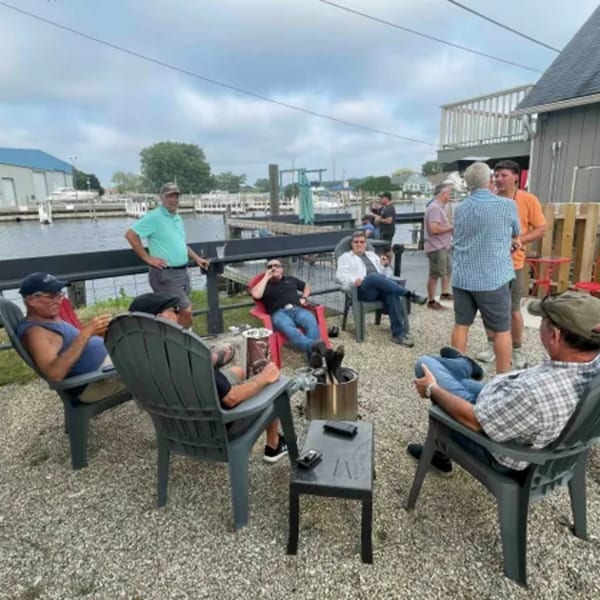 Men relaxing in chairs on a gravel patio by the water at On the River Tavern & Mo’s Cigar Bar.