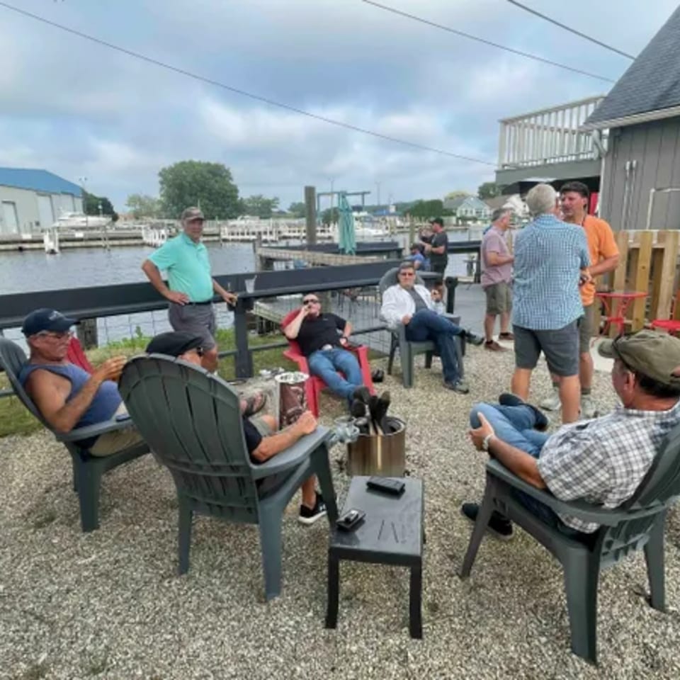 Men relaxing in chairs on a gravel patio by the water at On the River Tavern & Mo’s Cigar Bar.